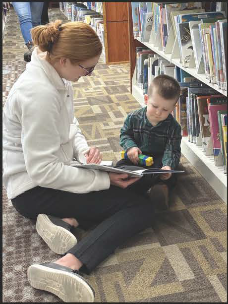 Adult reading to a child on the floor in the children's section of the Library.