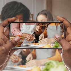 Two women cooking together with a third person recording it on their phone