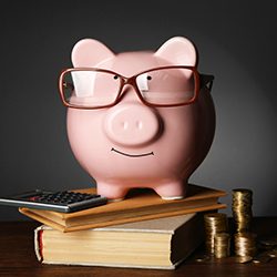 A piggy bank with glasses standing on a stack of books with a calculator and stack of coins.
